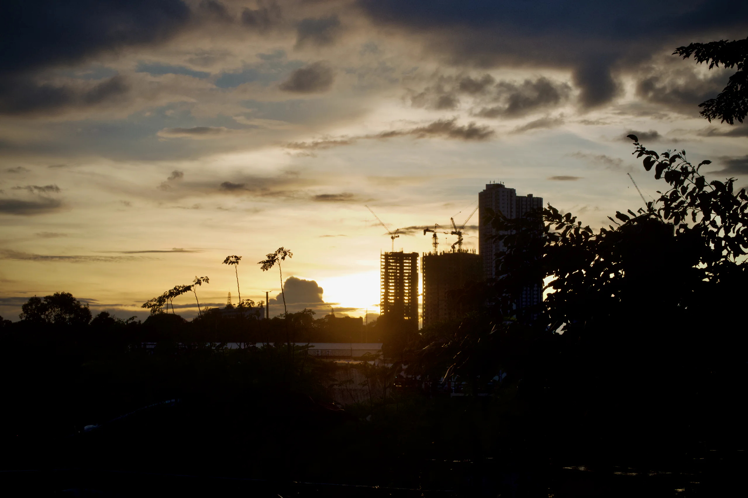 A dramatic sunset glows behind silhouetted trees, cranes, and high-rise buildings, while dark clouds stretch across the evening sky.