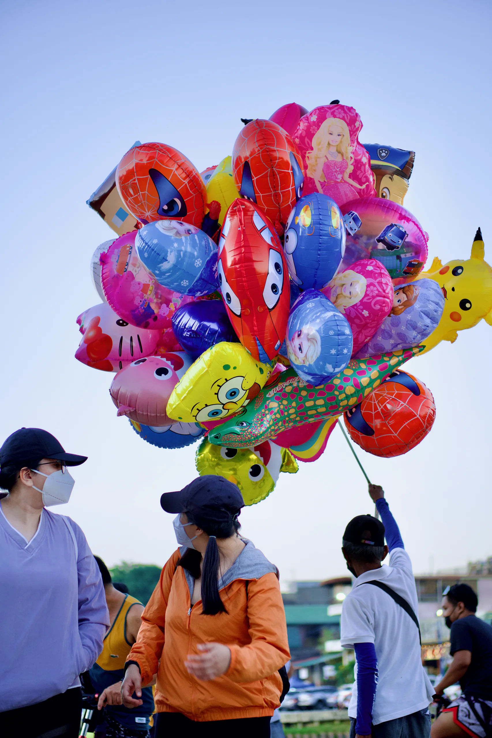 A balloon vendor holds a large cluster of brightly colored cartoon-character balloons above a group of pedestrians. The balloons fill most of the frame against a pale blue evening sky.